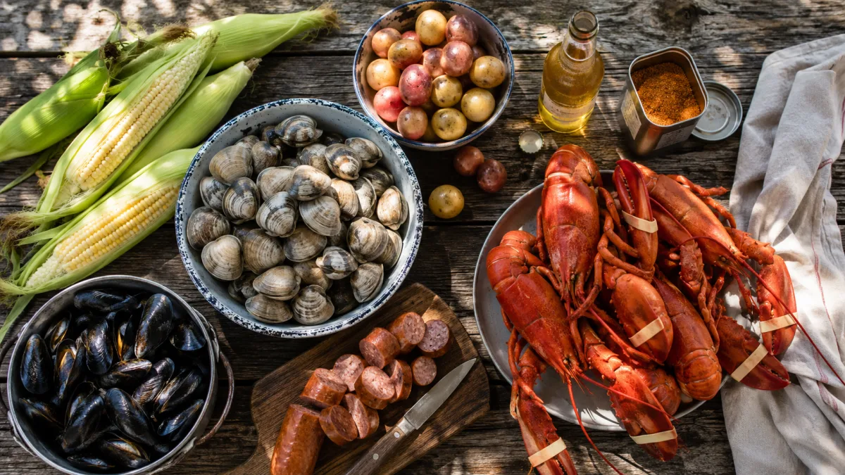 Fresh clambake ingredients arranged on a weathered wooden table — clams, mussels, lobster, corn, and sausage